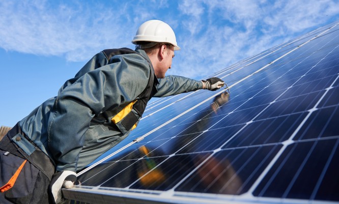 Side view snapshot of workman, wearing uniform, working gloves and helmet, setting a shiny new solar battery with help of hex key, blue sky on background. Green energy concept